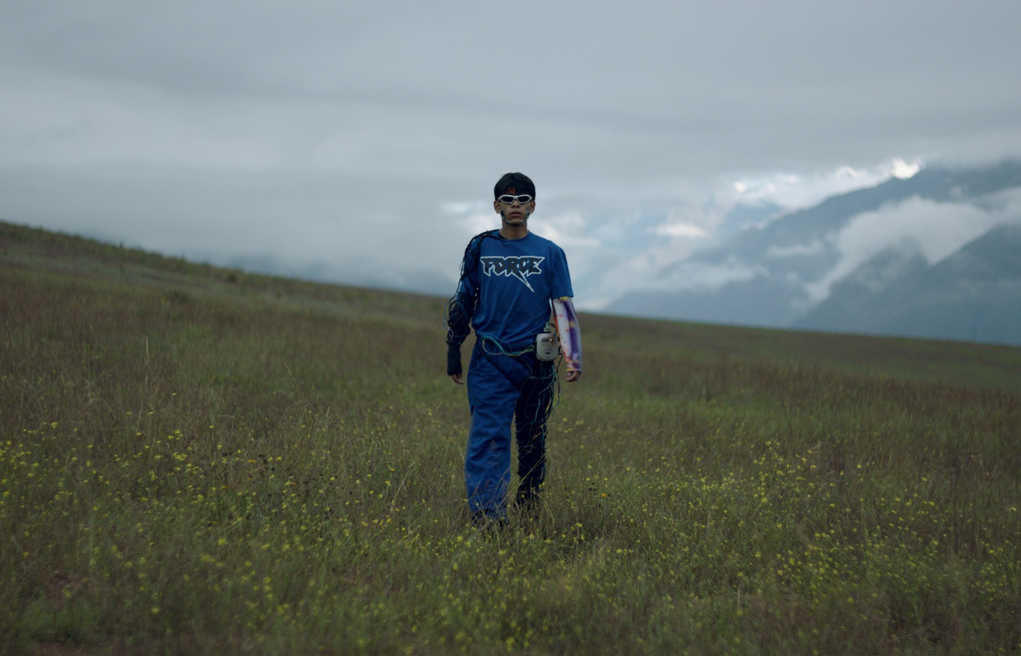 Still de 4EBER: Eber camina por un campo de flores amarillas con cables y un traje azul, con los Andes nublados al fondo